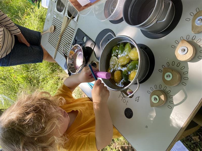 a young boy scraping chalk into a pan on the mud kitchen.