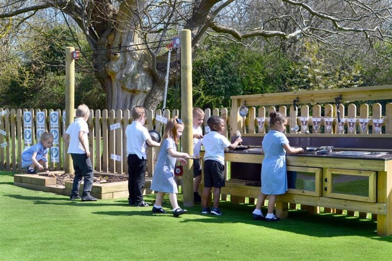 children in white, light blue and black school uniform stand and play with the mud kitchen and the rope and pulley mover and the sand box