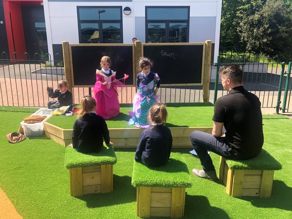 Two little girls are stood on an outdoor Performance stage as they wear princess dresses. Two children and an adult are sat on artificial grass topped seats, watching the performance.