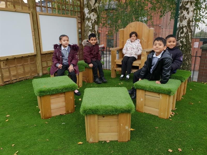 children pose on the story telling chair and on the grass topped seats facing the camera and smiling