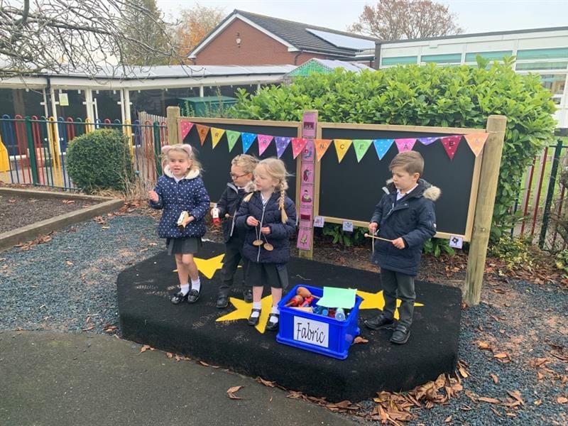 4 children are on the Outdoor Early Years performance stage, which was designed and constructed by Pentagon Play. The children are using different materials from a box labelled "fabric" to carry out there play.