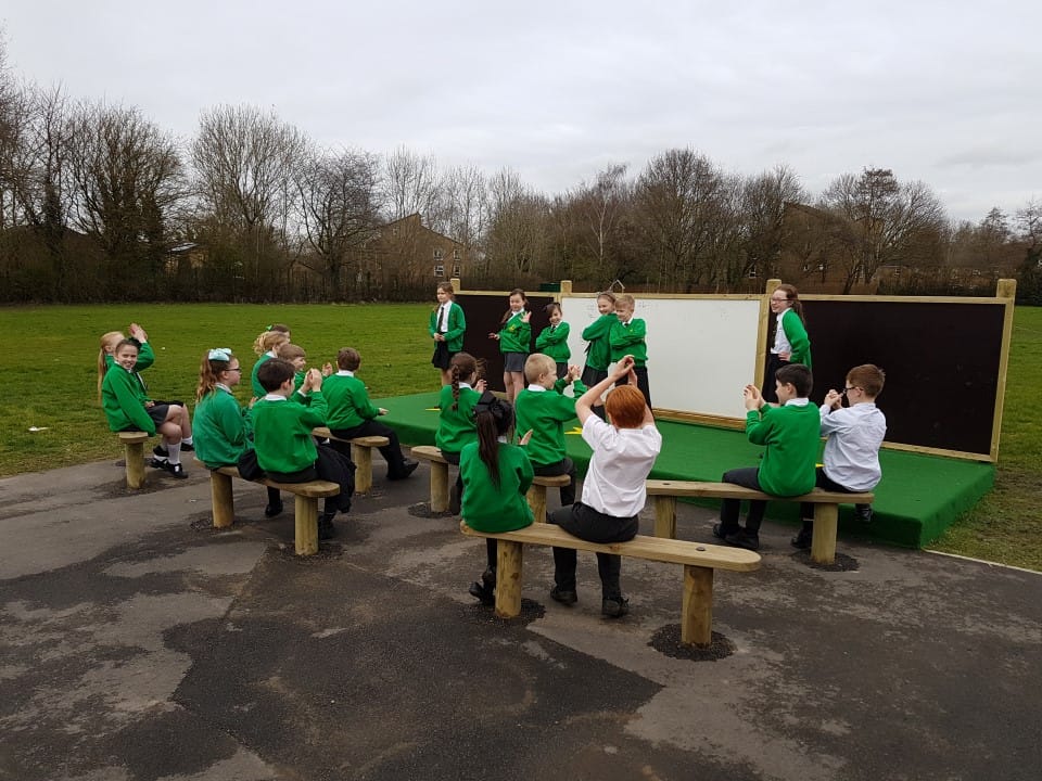 A small group of children are posing on a stage as a large group of children who are sat opposite the stage are clapping.