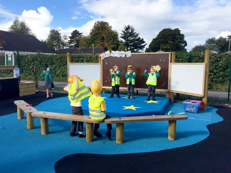 3 children are dressed up as builders as they perform a song on an outdoor stage, created by Pentagon Play. 2 children are sat on wooden benches as they watch the show, also dressed up as builders. 1 child can be seen to the right of the stage dancing.