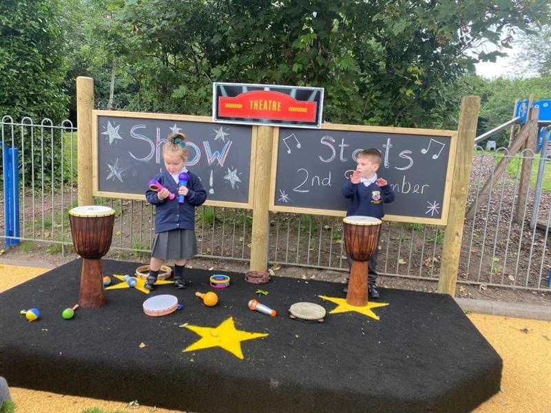 2 children are stood on our outdoor early years development performance stage. There is different musical instruments scattered around the stage and the children are stood next to a conga drum and are ready to perform for their talent show.