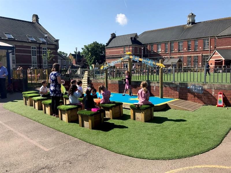 a little girl dances and performs on the performance stage for her classmates who sit and watch from the artificial grass topped seats on a sunny day