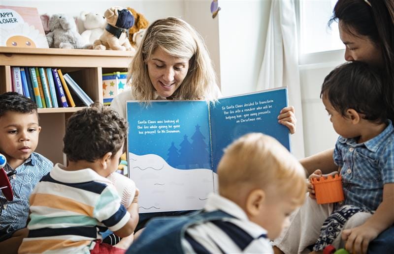 children sat around in the classroom whilst a teacher is showing them a page of the book.