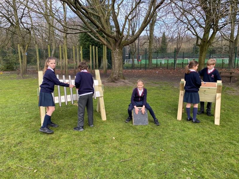 children sit on the drum seats in the music pack whilst others stand at the tongue drum and the chimes and play the instruments