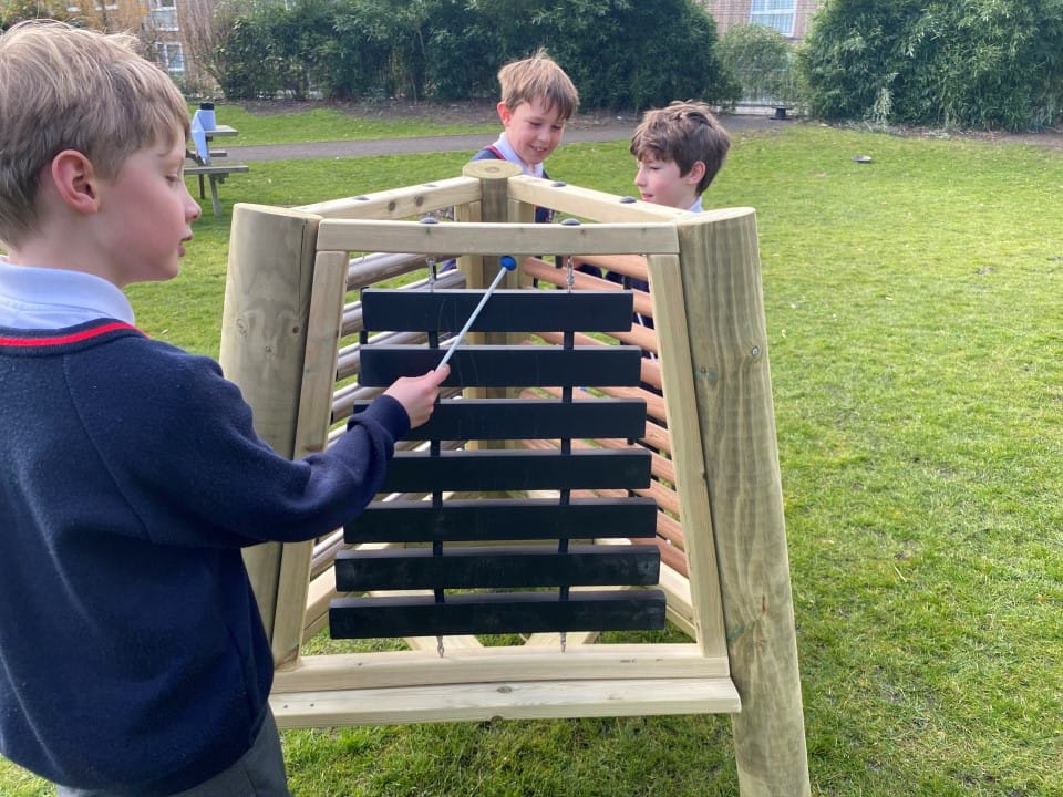 Three boys are playing with a freestanding music easel, talking to each other and smiling.