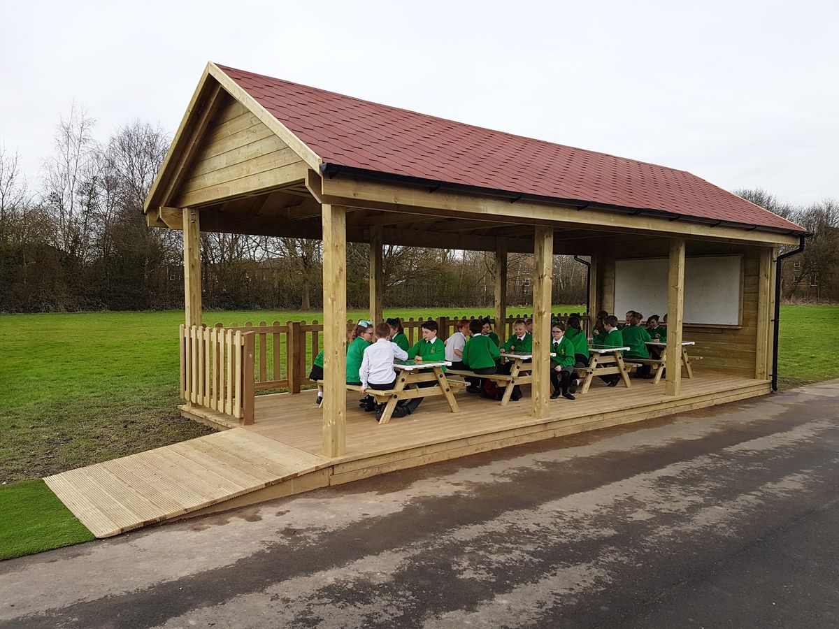 A large outdoor classroom with numerous school picnic benches. Children are sat at these benches as they talk.