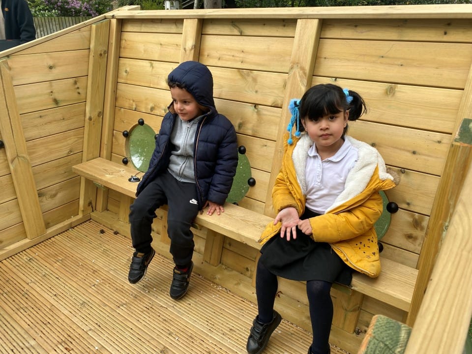 Two children are sat on a wooden bench that is inside a Play Ship.