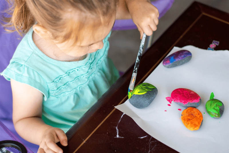 a cild sits with a paint bursh and paints a stone