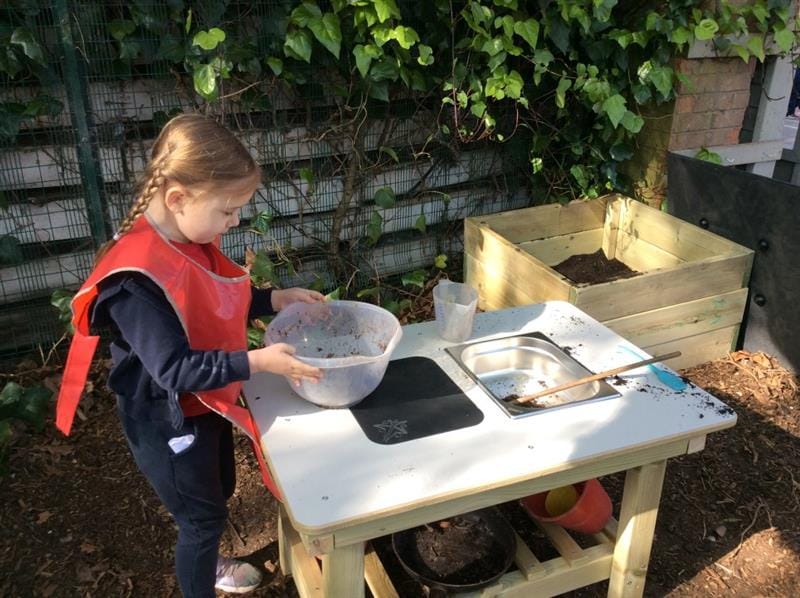 a little girl stands by the mud kitchen and mixes mud together in a bowl