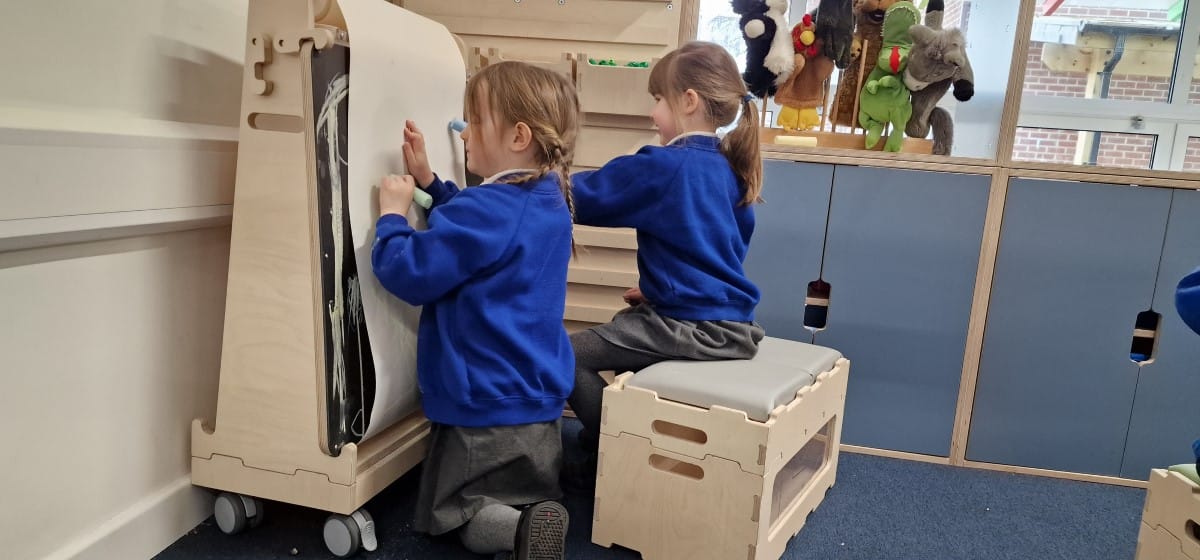 Two girls are holding sticks of coloured chalk and are drawing on a piece of paper that has been attached to the chalkboard easel.