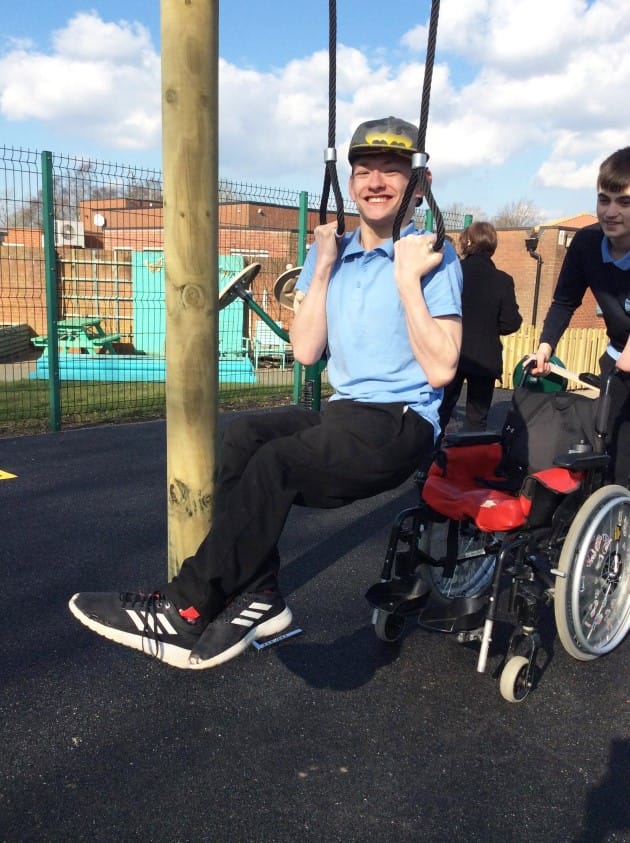 A boy is holding himself up on two pieces of rope by his hands, as another boy is holding a wheelchair behind the boy holding the ropes.
