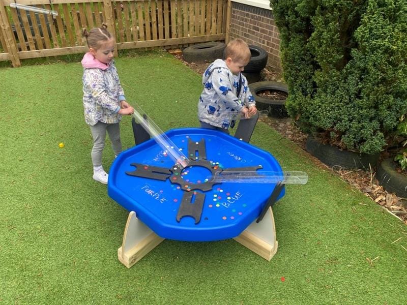 two children stand aroudn the blue tuff spot table and play with counters down the water channel funnels