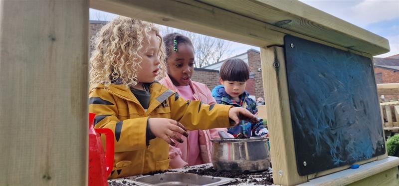 three children stand and play together on the essentials mud kitchen