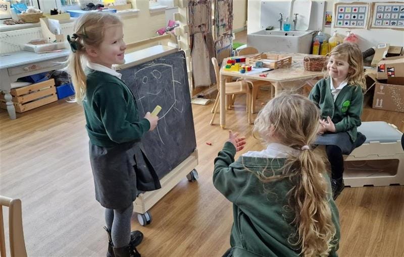a child stands at the indoor art easel and draws a face and shows it to her friends