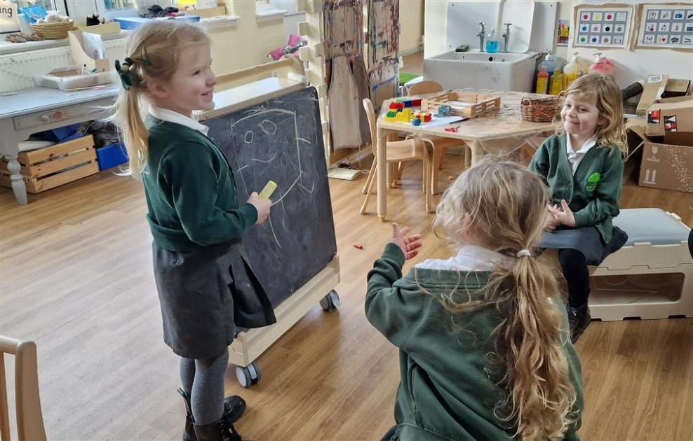 A little girl is drawing on a chalkboard and is presenting her work to two other little girls.