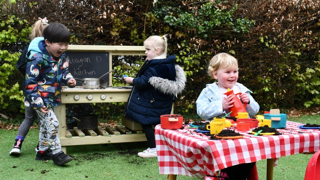Three children are playing with a Mud Kitchen and are cooking up something, whilst another child is sat at a table that has been decorated to look like they're at a restaurant, with a variety of mud dishes placed on it.