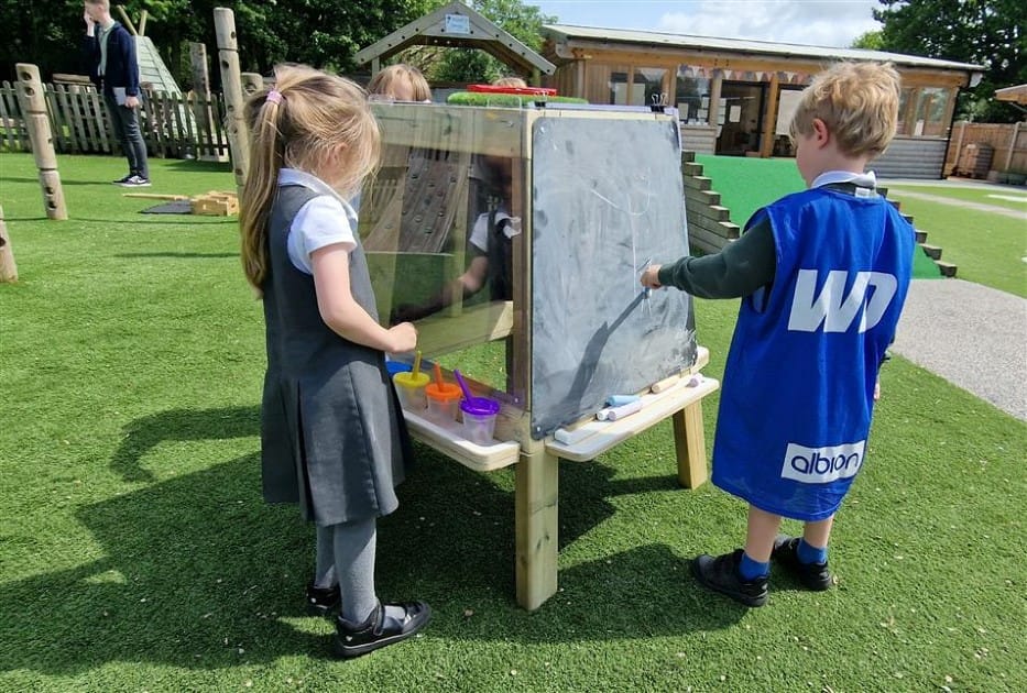 Three children are engaging with the group art easel. A boy is wearing a blue jacket as he draws on the chalkboard, whilst the little girl to his left is looking at all the paints she can use. The other child is at the back of the easel, drawing on a chalkboard. The group art easel is set up on an artificial grass top.
