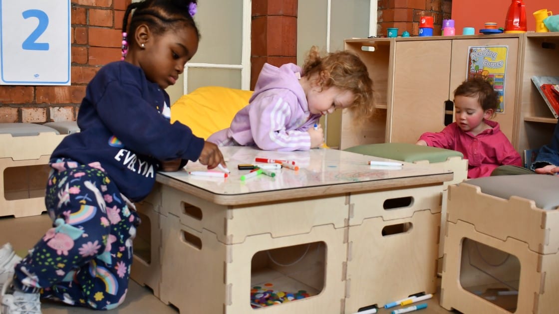 Three children are using different coloured whiteboard markers to draw on the whiteboard surface that is provided from our Stack and Sit Package. The classroom is in an EYFS environment, with the three children looking very young.