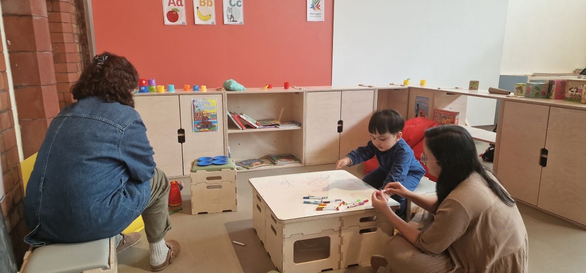A group of children are sat around a table and are taking part in an activity with the help of teachers/assistant teachers.