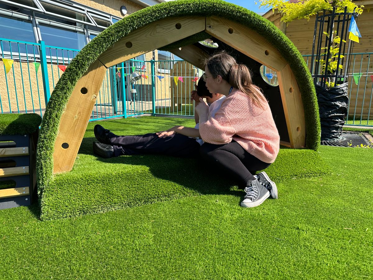 A child with EBSA is sat within a grass tunnel reading a book as a teacher is sat beside them and helping them.