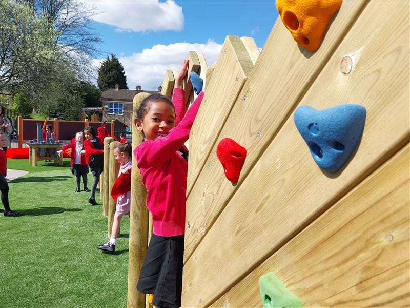 A young girl climbing on the climbing wall as she looks towards the camera and smiles.
