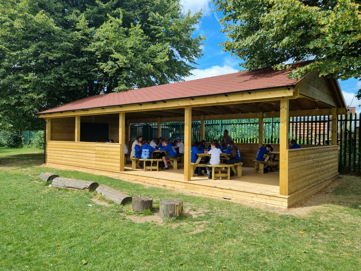 A large group of children are sat in a freestanding canopy and are all around tables. A teacher can be seen in the middle of the structure.