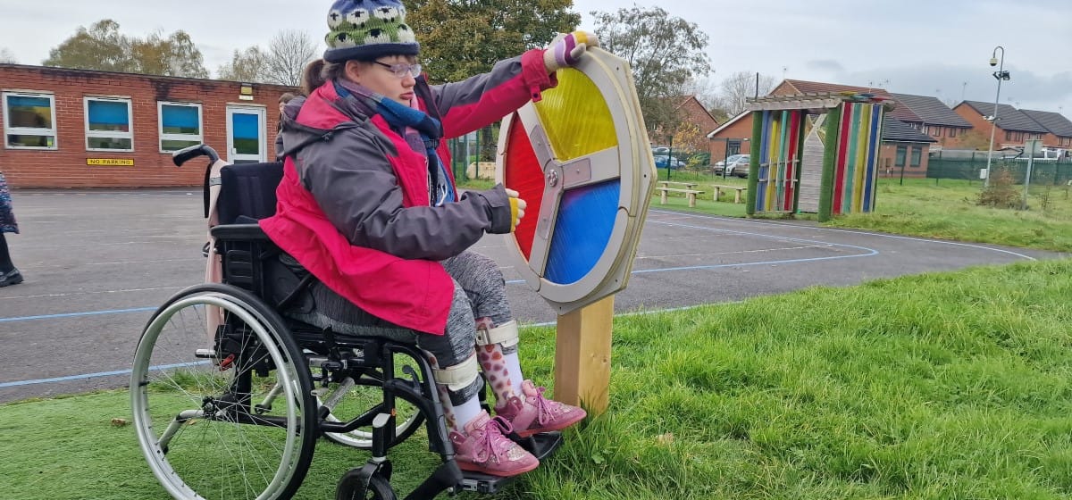 A girl in a wheelchair is playing with a sensory spinner that has been installed on grass.