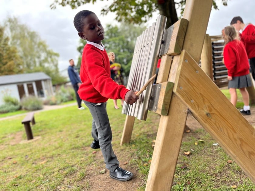 A child is playing with the freestanding chimes in a grass area. The child is using a stick to play the chimes as the other children engage with the other playground equipment surrounding him.