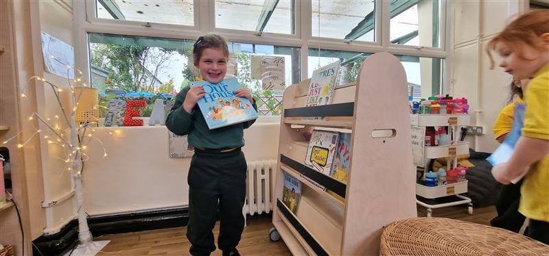 a little girl holds a book from the bookcase on wheels and poses with it as another child is holding a book selected from the bookcase