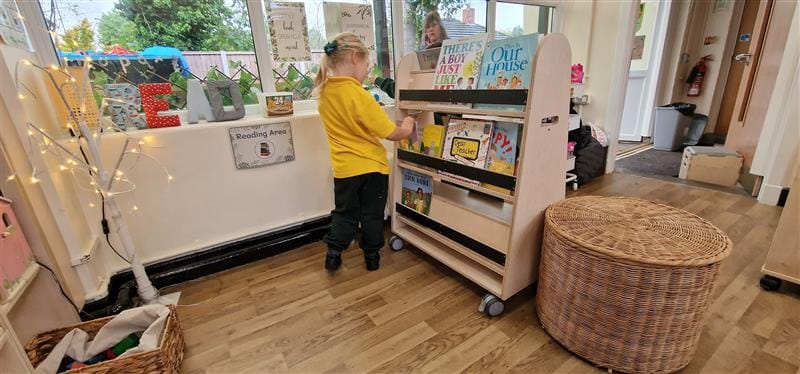 a little girl in a yellow top picks books from the bookcase on wheels within a classroom setting.
