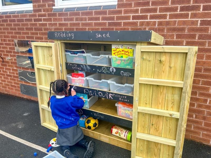 a little girl kneels on the floor in front of the sycamore store in order to reach the resources within the store cupboard
