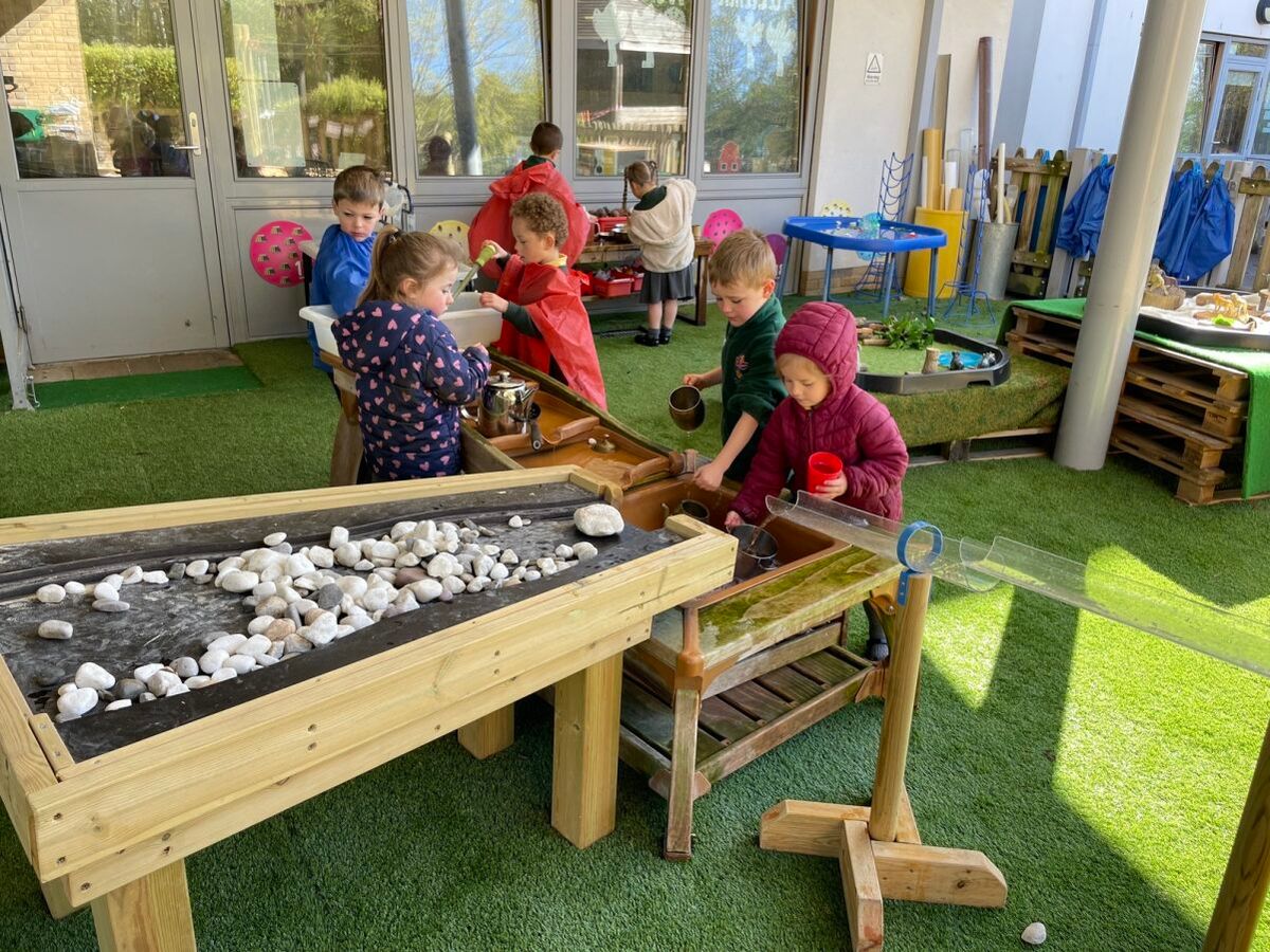 A group of five young children playing with the Pentagon Play Dam. They are collecting water from a pipe and pouring it down the dam to see how the water reacts.