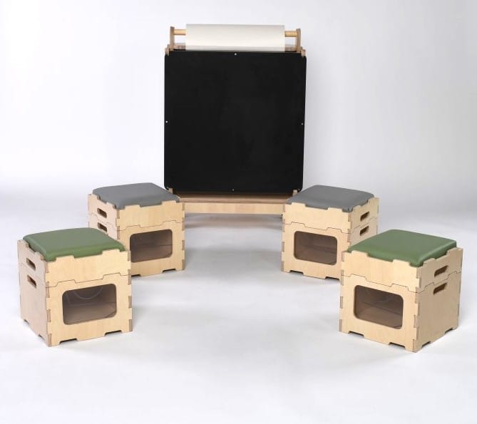4 stack and sit blocks are placed in front of Pentagon Play's indoor art easel. 2 of the Stack and Sit blocks have a sage green seat cushion, where the other 2 have a grey cushion