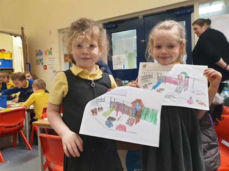 Two girls are holding up their drawings of playgrounds to the camera.