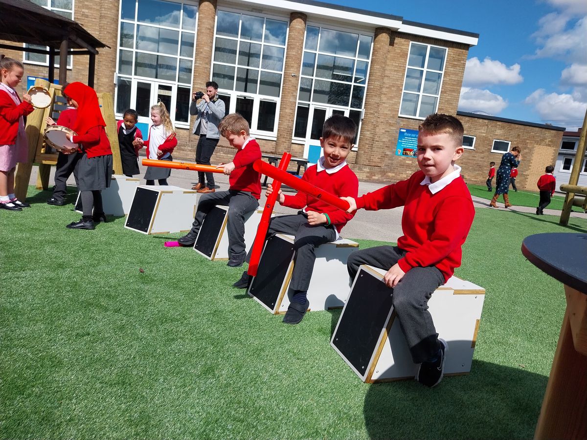 8 children wearing red school uniforms are playing with Pentagon Play's outdoor musical instruments, including drum seats, a music easel and tambourines. All the children look happy and excited to be playing with the equpment.