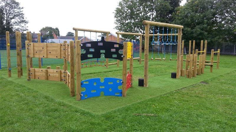 Playturf being placed underneath a trim trail on a playground. The playground equipment is made out of wood and plastic, surrounded by trees and a blue fence.