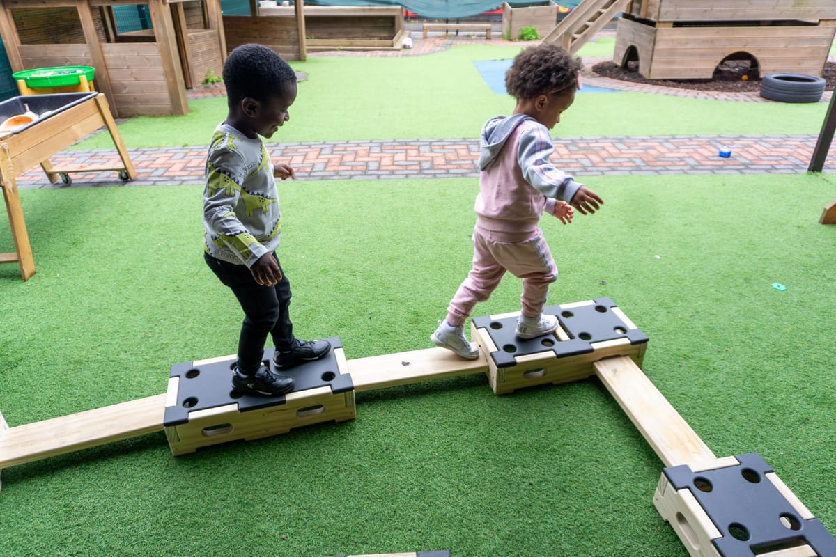 Two children are playing on the Play Builder Engineer Set and are balancing across a wooden beam. They are in a single file line.