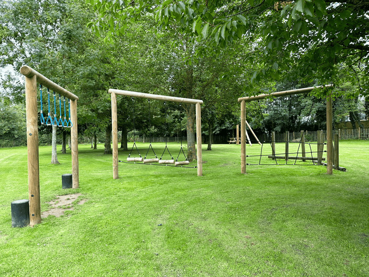 3 parts of a trim trail have been installed on grass. The first part has rope handles to swing across. The second has a log traversal step and the third one has a rope traversal element.