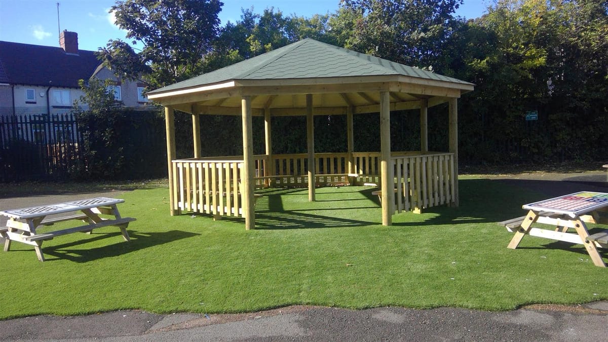 A gazebo outdoor classroom that has been installed on artificial grass, with two picnic benches beside it.