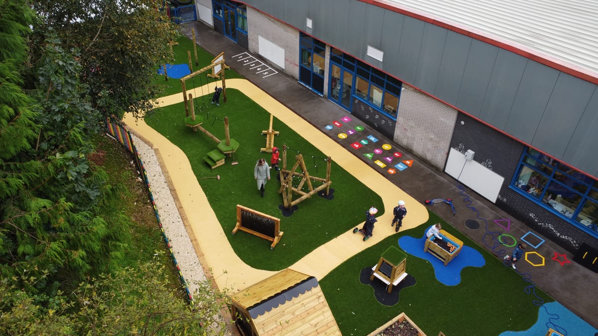 A birdseye view of a playground with artificial grass surfacing and a wetpour road design installed on top. Children can be seen playing with a variety of play equipment on the surface