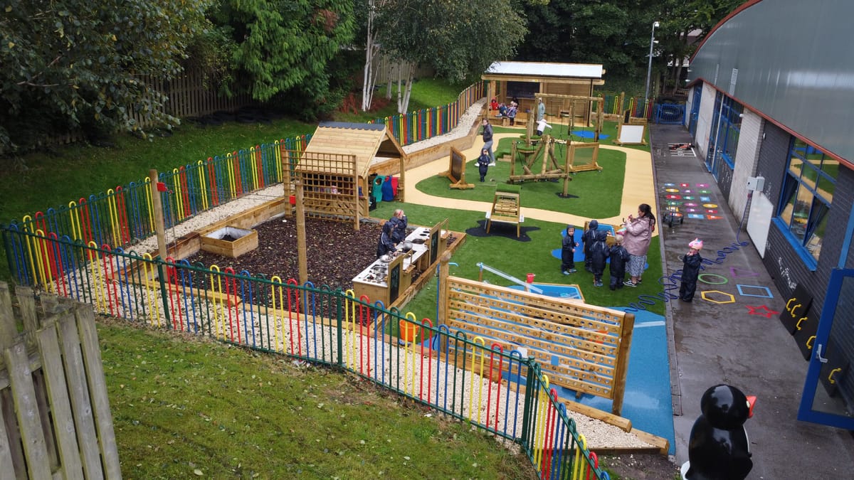 An EYFS play area that is surrounded by colourful fencing. Within the fenced area. a variety of play equipment can be spotted with children exploring the area.