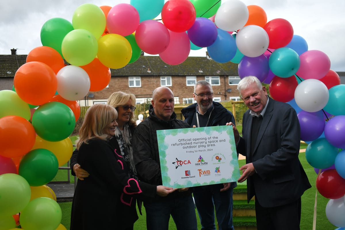 A group of men and women are holding up a card which states "Official opening of the refurbished nursery space and outdoor play area. Friday 1st March 2024". They are stood underneath a multicoloured balloon arch.