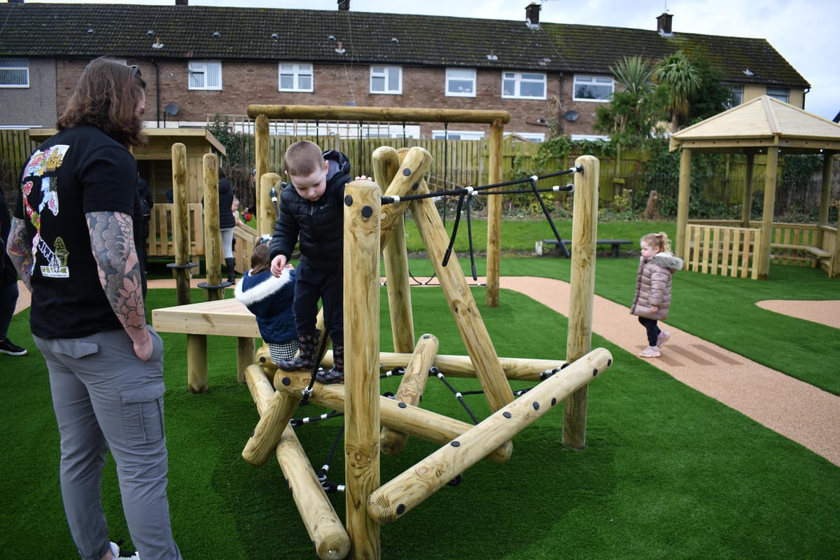 A man is stood beside his child as the little boy explores a climbing frame on a playground.