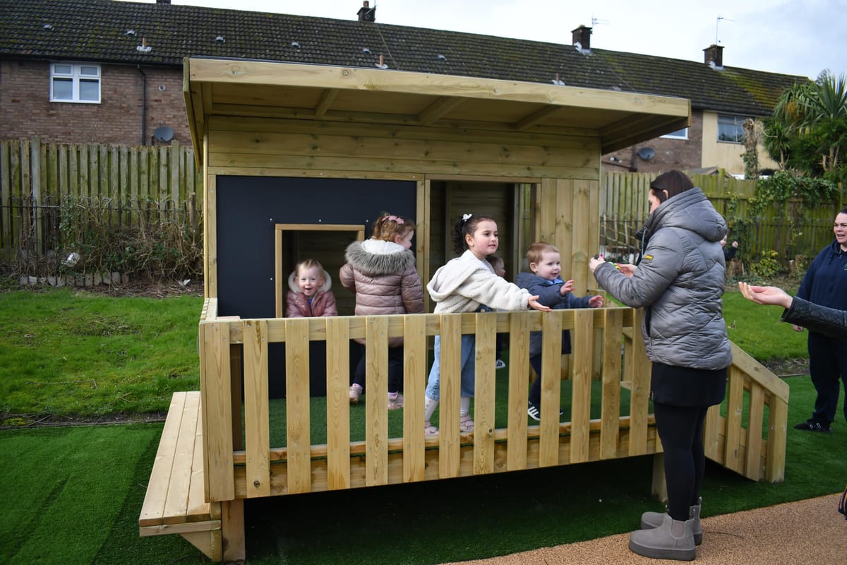 A small group of children are stood on a small wooden balcony and are looking at a teacher. The teacher is making eye contact with them and is performing an action, which the children are mirroring.