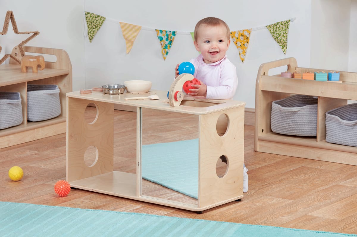 A toddler is playing with a simple role play kitchen set, and is holding the tap. The baby is looking off-camera and smiling.