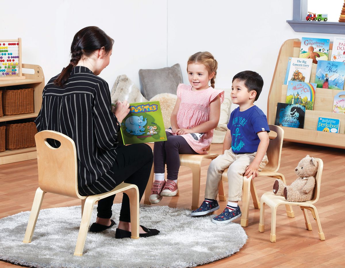 A teacher is sat on a teaching chair as she is showing two children the pages of a book. Two children are sat in front of her on smaller wooden chairs, with a teddy being sat on a small chair. The children are looking into the book.