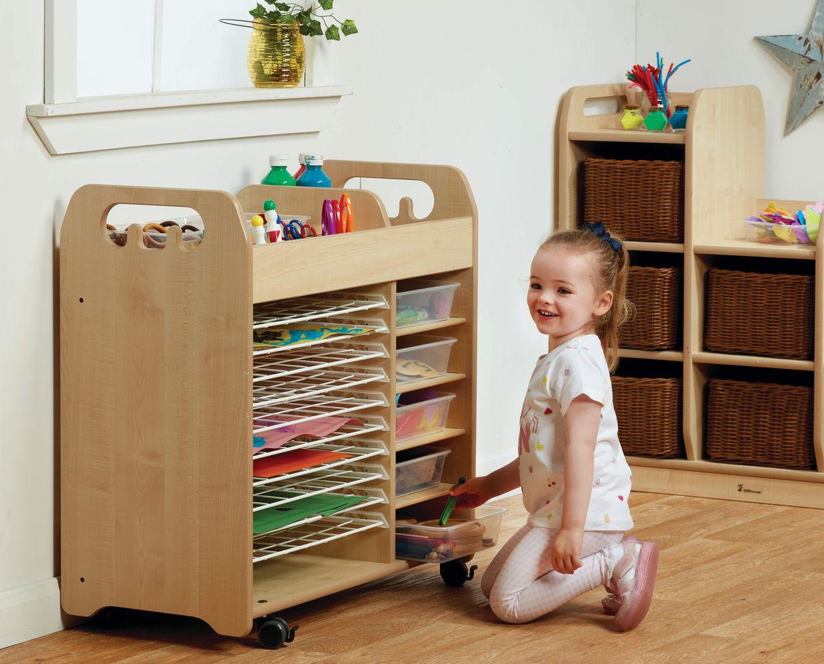 A little girl is kneeling and looking at the camera, smiling. She is currently looking through a variety of different trays that are placed inside a tray storage unit. Each tray is holding a different arts and crafts material.